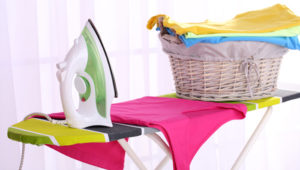 Basket with laundry and ironing board on light home interior background